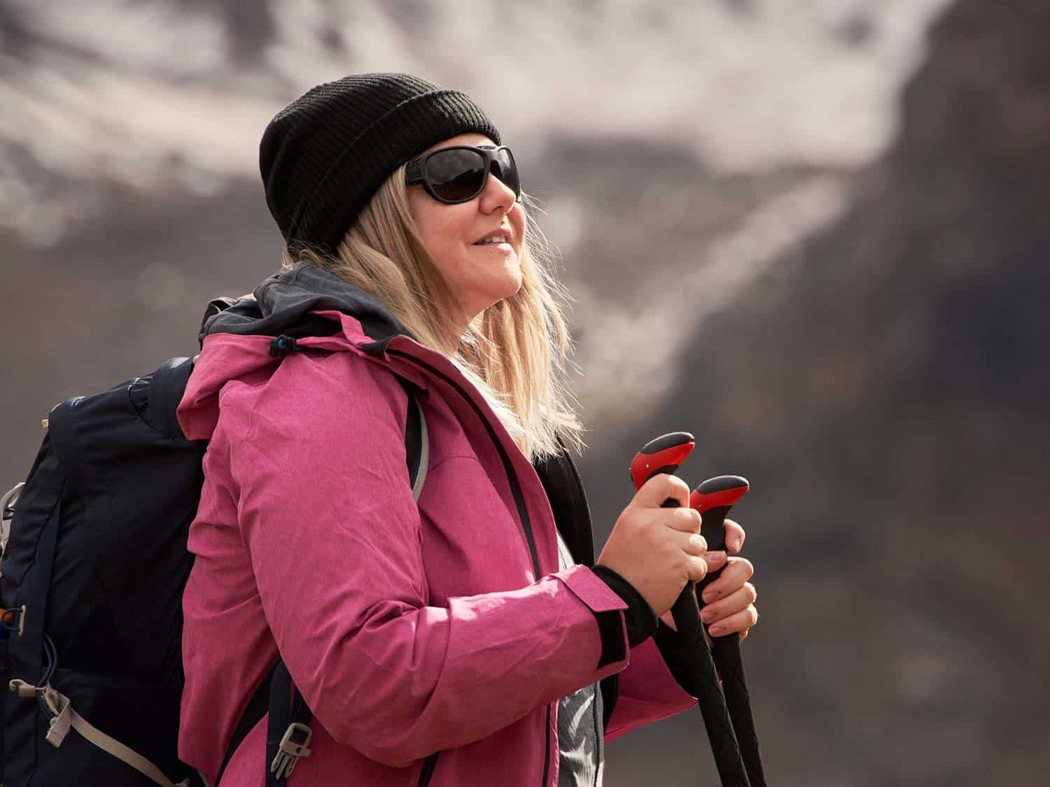 Person stood in pink coat, blue backpack, black hat and sunglasses using hiking poles with out of focus mountain scene in background
