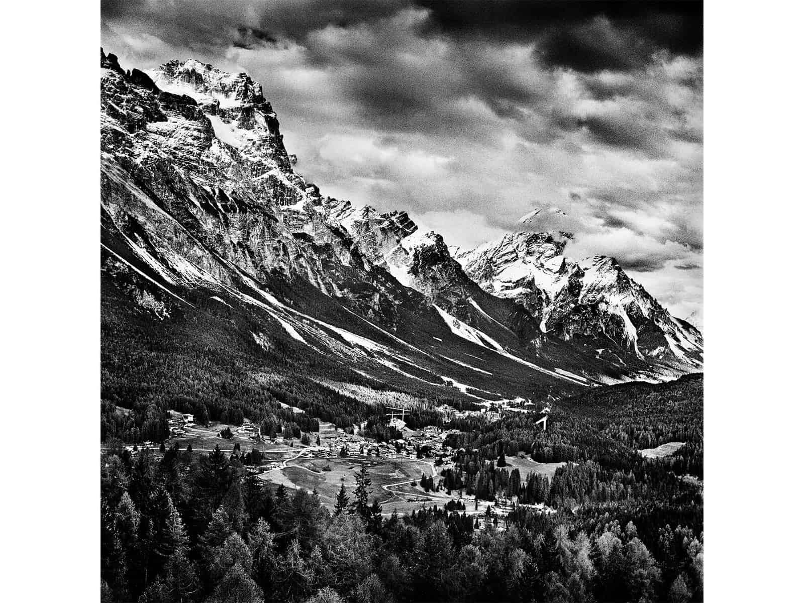ID: A square, black and white image. The contrast is very high in this mountain image. In the foreground are alpine mountains that are snow capped. In the background there are more snow capped mountains. The clouds are dramatic in the sky.