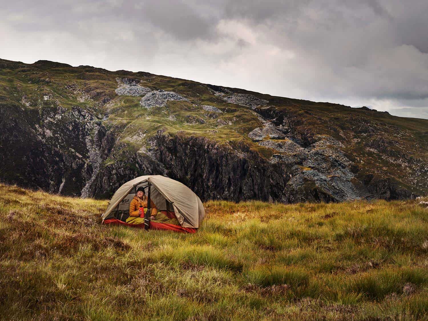 person in yellow coat sits in Near Zero Ultralight backpacking tent in mountainous background