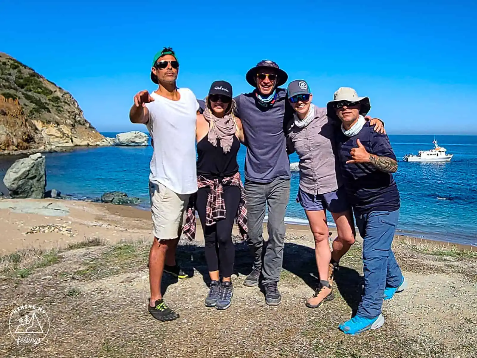 ID: A landscape image. Five people stand close together taking a photograph, they look happy. The are all wearing warm weather clothes such as shorts and t-shirts and hats. There is a bright blue sky behind them and they are stood next to a cove wit…